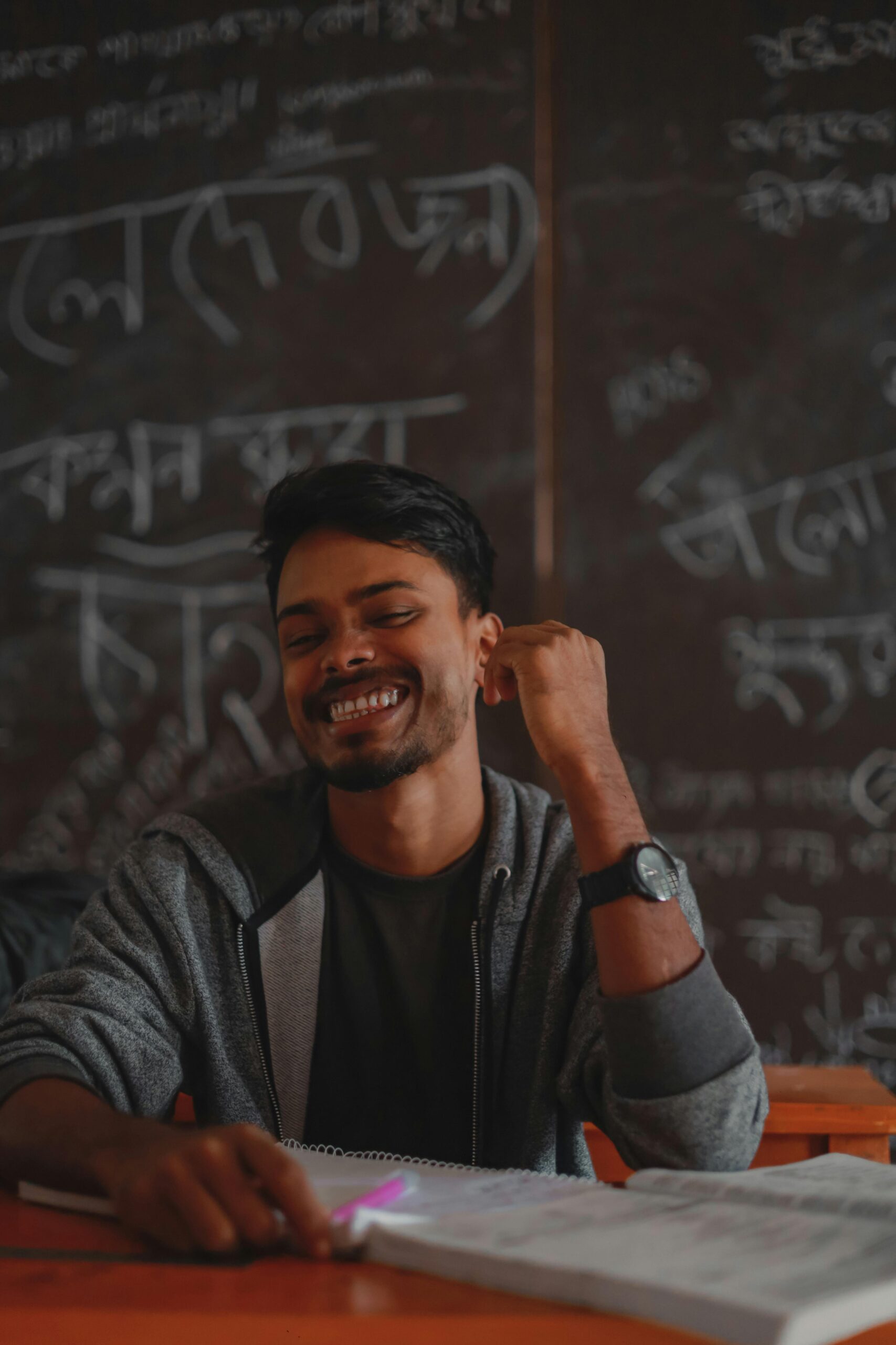 Young South Asian student smiling in front of a chalkboard in a Bangladeshi classroom.